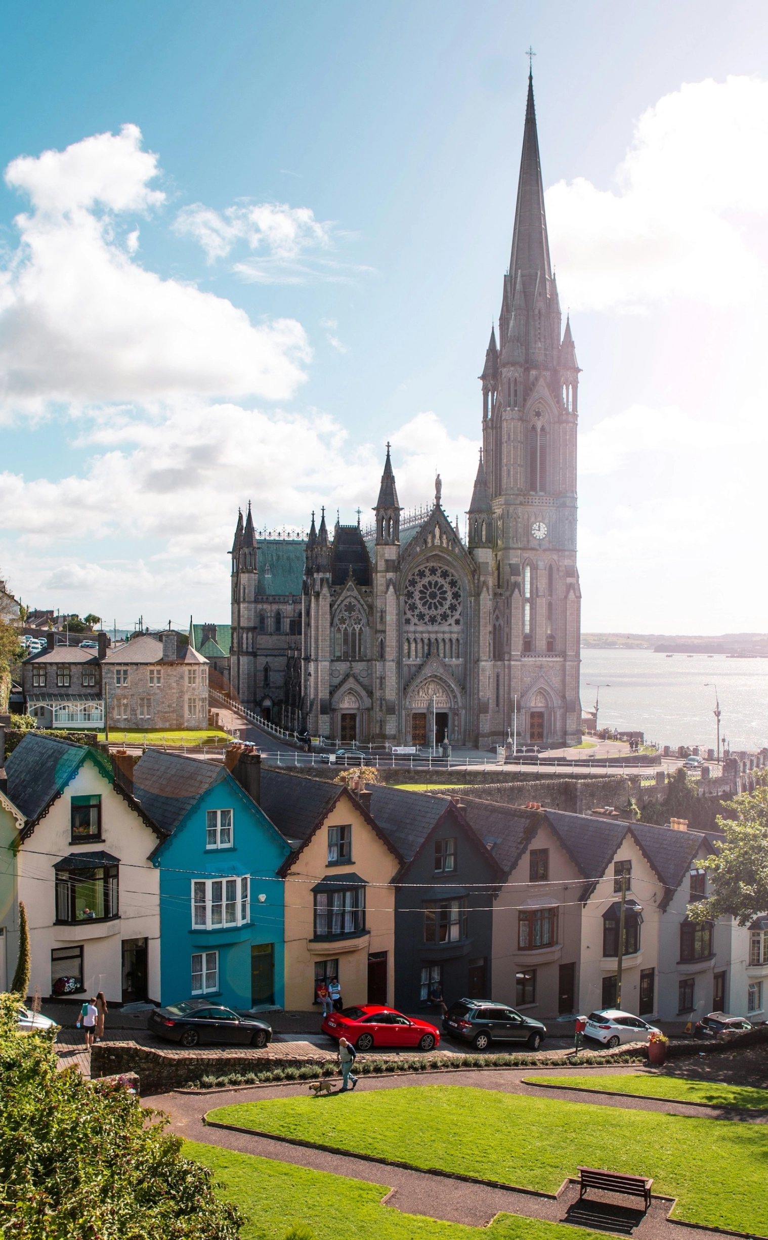 An aerial view shows the majestic grey St. Colman's Cathedral with its tall spire overlooking a harbor, with a row of brightly painted terraced houses in the foreground and a green park with a pathway.