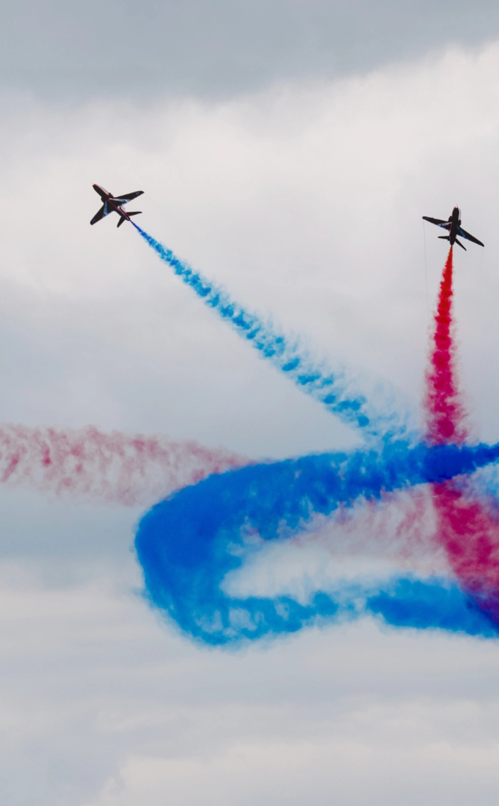 Two dark-colored jet planes fly through a cloudy sky, leaving prominent trails of red and blue smoke that intersect and curl in an aerial display.