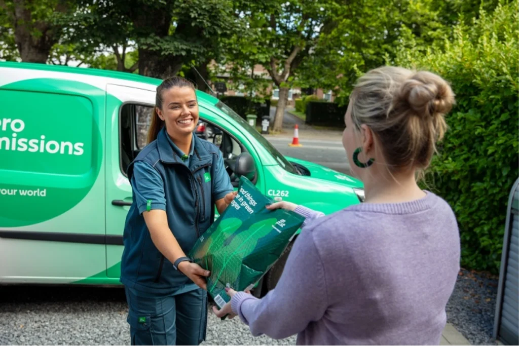 A smiling postal worker in a green uniform hands a large green package to a customer, with a bright green 'Zero Emissions' delivery van parked behind her.