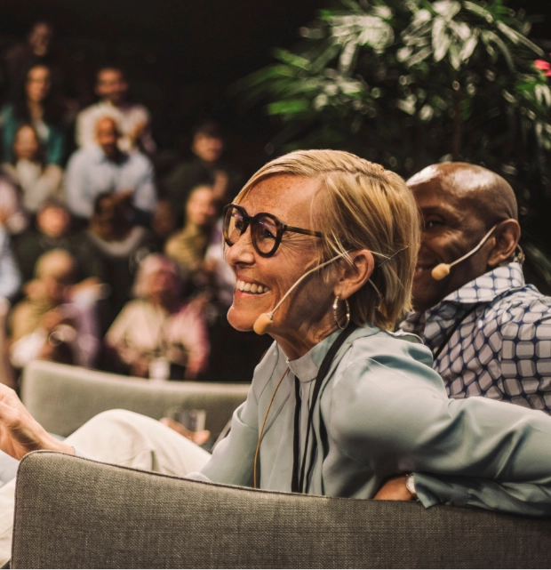 Two smiling speakers with headsets engaged in a panel discussion at a conference with audience in background