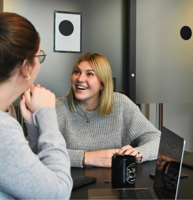 Two professional women smiling and talking in a modern office meeting room with laptops and coffee